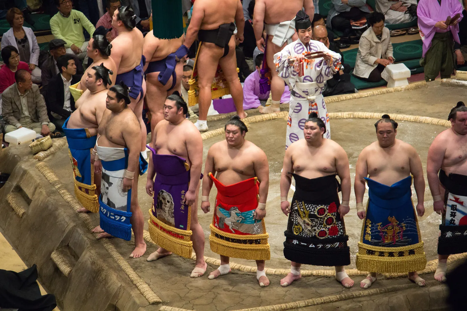 A ring of sumo wrestlers in colourful ceremonial aprons standing on a clay platform in an arena, with the crowd around them