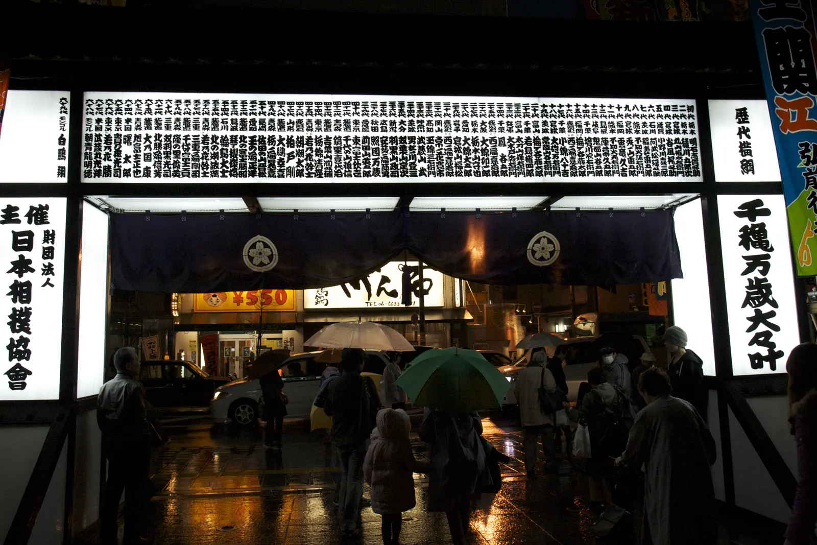 The lit entrance of a sumo venue at night, with tall banners of black calligraphy on white and people standing under umbrellas
