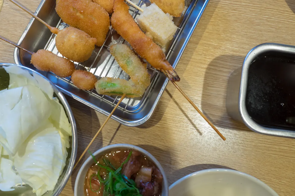 A metal tray holding golden breaded and deep-fried skewers beside a bowl of shredded cabbage and a small dish of dark sauce