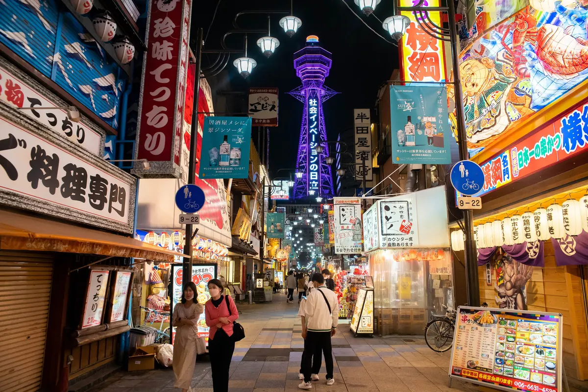 A tall lit lattice tower glowing purple above a street of neon signs and lanterns in Shinsekai at night