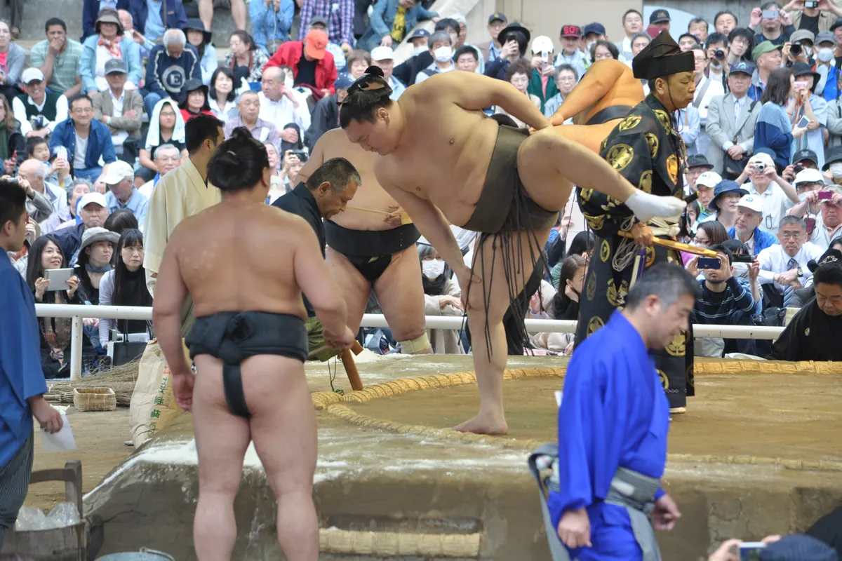 A sumo wrestler balanced on one leg with the other raised high beside the edge of a ring, in front of a crowd