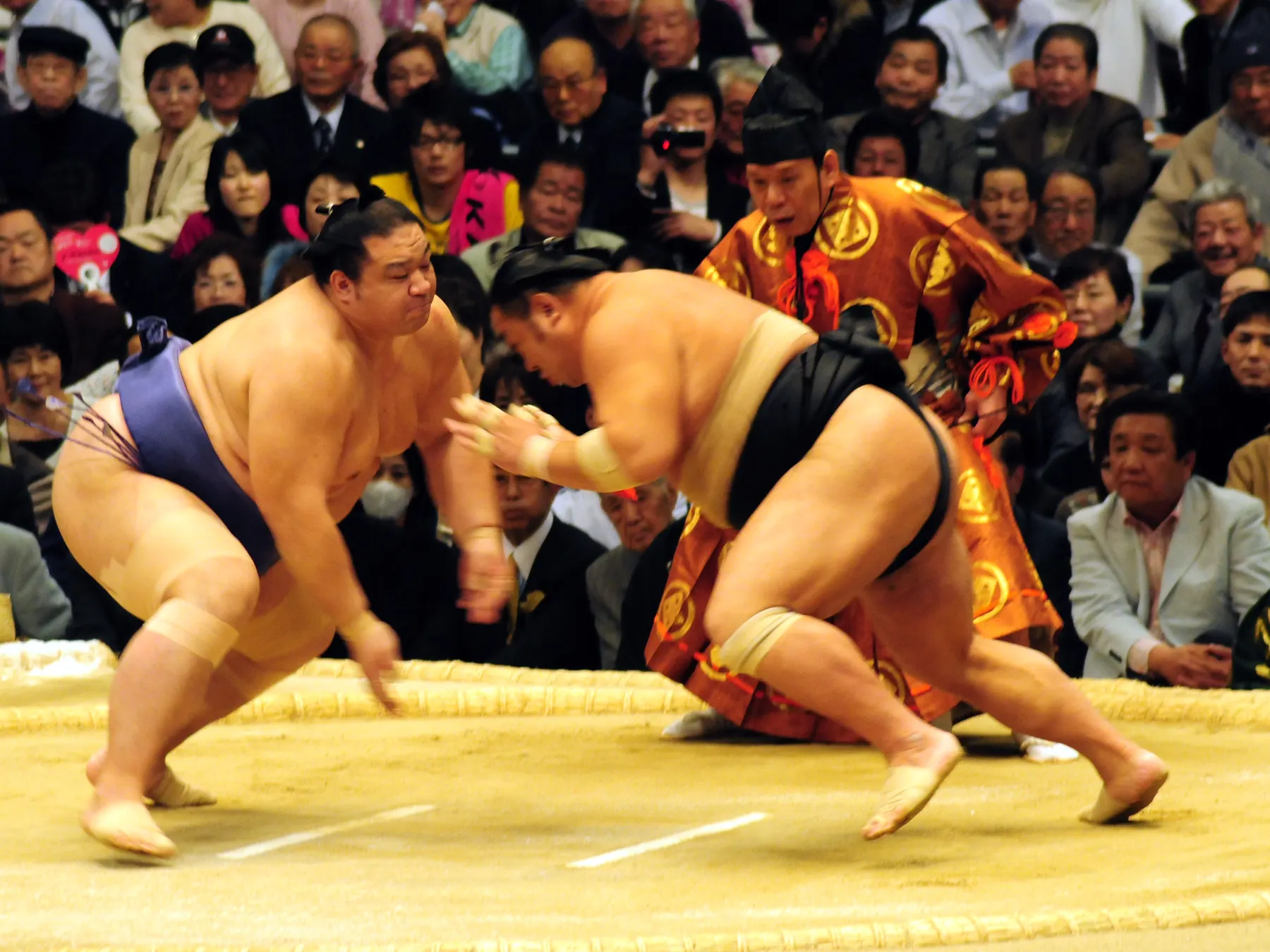 Two sumo wrestlers charging at each other at the start of a bout in a straw-bale ring, with a referee in orange ceremonial dress behind them and a seated crowd beyond